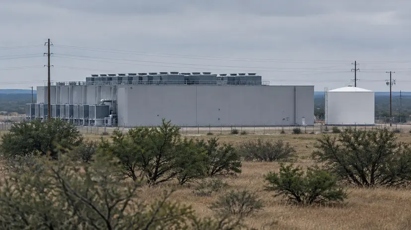 A windowless data center with rooftop cooling units and a water tank behind a fence on Texas brushland.