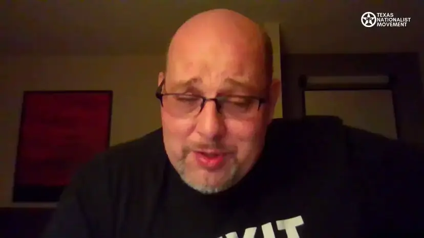 A bald man in glasses and a TEXIT shirt speaks on a video stream from a hotel room.