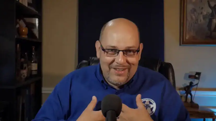 Daniel Miller smiles and gestures toward himself during a livestream, wearing a blue TNM shirt.