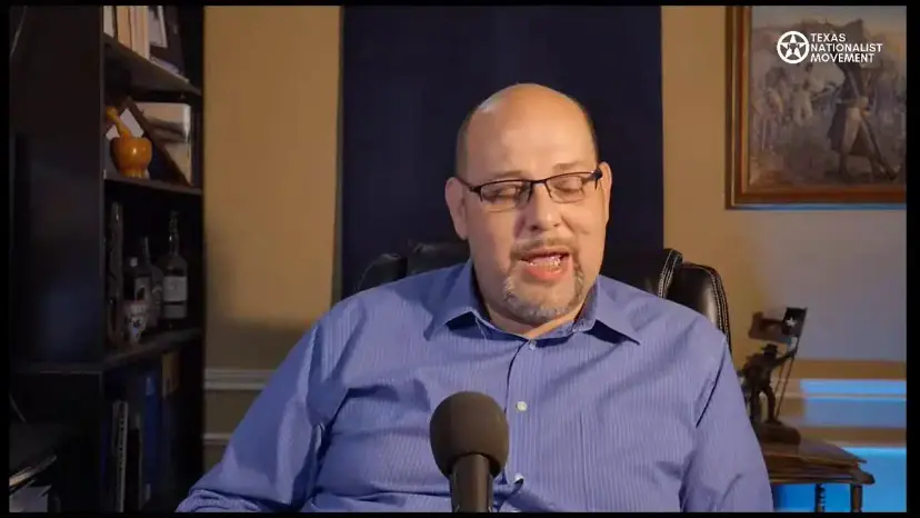A bald man in glasses and a blue shirt speaks into a microphone during a livestream.