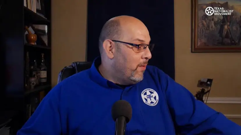 A bald man in a blue TNM shirt looks to the side while sitting at a microphone.