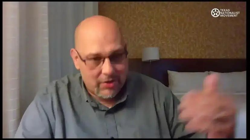 A bald man in glasses gestures while speaking on a video stream from a hotel room.