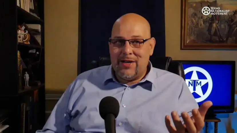 Daniel Miller speaking to the camera from a home studio with a Texas Nationalist Movement logo on a screen.
