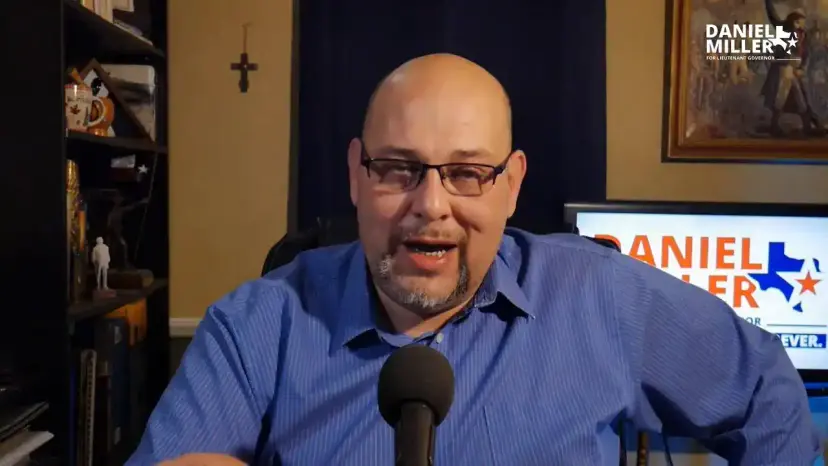Daniel Miller speaks at a microphone in his office with a campaign sign behind him.