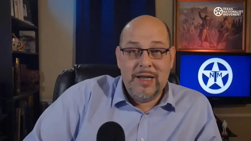 A bald man with glasses speaks to the camera in a studio with a Texas Nationalist Movement logo behind him.