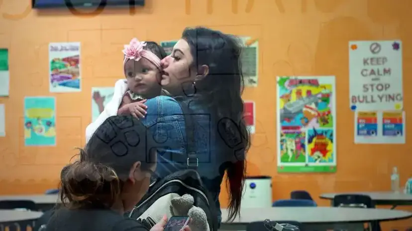 Young mother holding her baby girl in a classroom with colorful posters on an orange wall