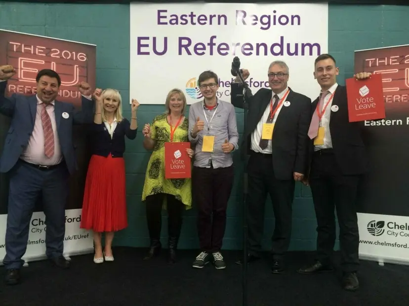 Six people cheer at the Eastern Region EU Referendum count, two of them holding 'Vote Leave' signs.