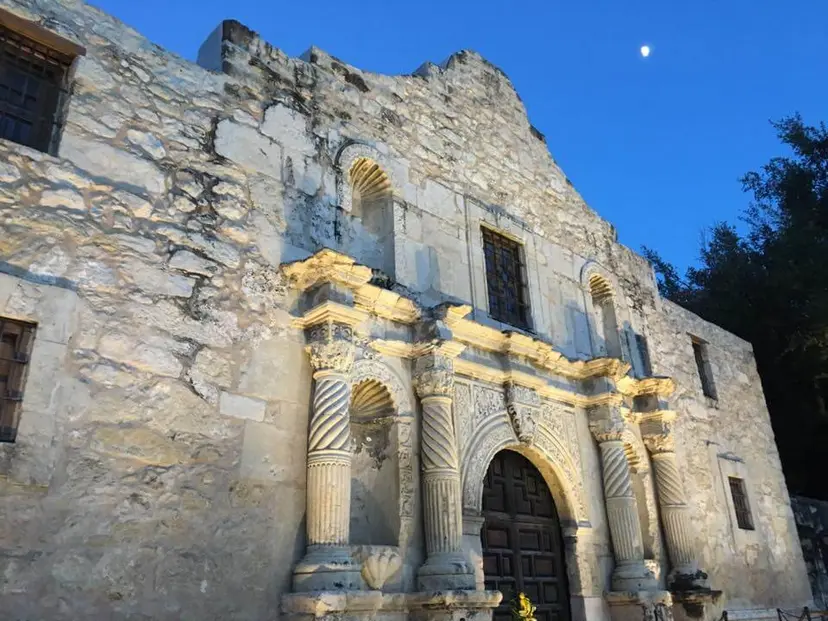 Stone facade of the Alamo church in San Antonio, lit by evening sun under a blue sky with the moon.