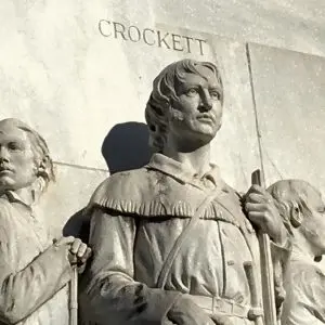 Carved stone figure of Davy Crockett beneath his name on a marble memorial, with other defenders beside him.