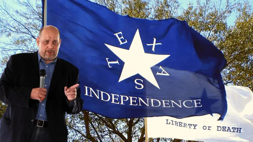 Daniel Miller speaking into a microphone in front of a blue flag reading Texas Independence.