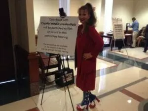 A woman in a red dress and star-patterned boots stands beside a sign about Capitol media credentials