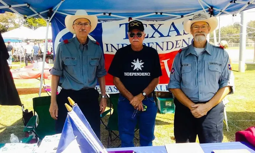 Three men, two in cowboy hats and uniforms, standing at a Texas independence booth at an outdoor pow wow.