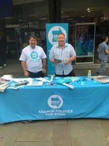 Two men staff a Brexit Party campaign stall with a blue table reading 'Change politics for good.'