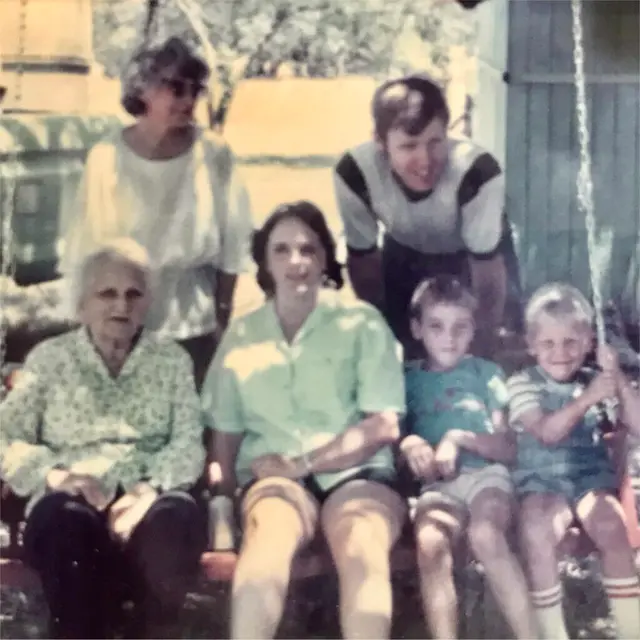 Faded family snapshot of an elderly woman, two women, a young man, and two small boys on a porch swing.