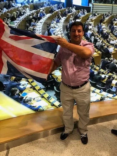 A man holds up a Union Jack flag inside a large parliamentary chamber with delegates seated below.