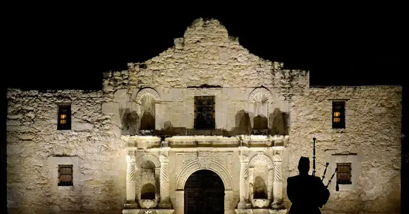 The Alamo church lit at night with a bagpiper in silhouette at the right.