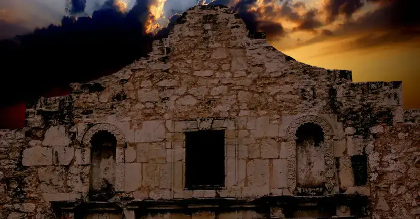 The stone facade of the Alamo church under dark, stormy clouds lit by an orange sunset.