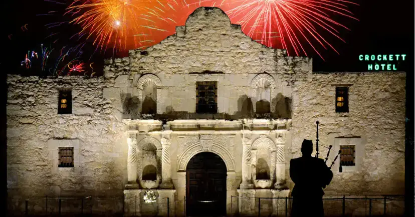 Fireworks bursting over the Alamo church at night as a bagpiper stands in silhouette.
