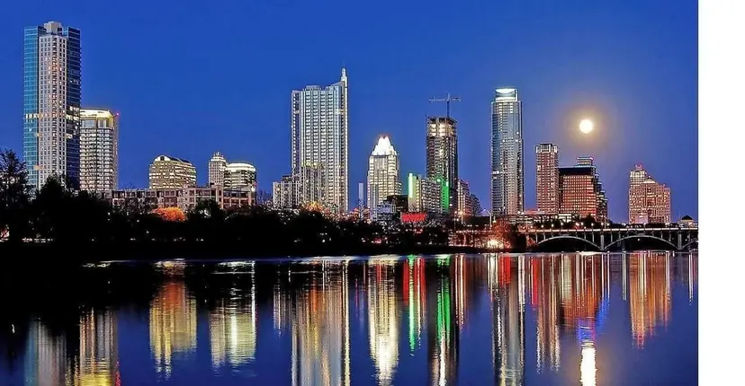 The downtown Austin skyline at night reflected in the river below a full moon.