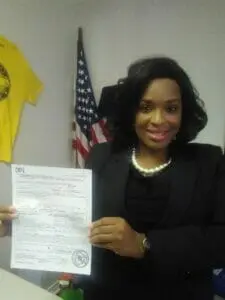 A woman in a black suit and pearls holding up a filed document beside a U.S. flag.