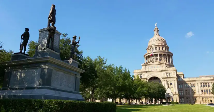Monument with bronze statues on the Texas Capitol grounds, with the Capitol dome under a blue sky.