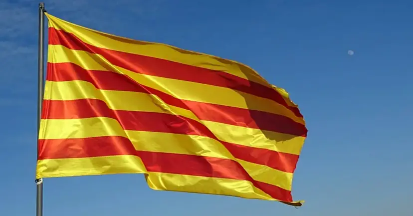 A red and yellow striped Catalan flag waving from a pole against a blue sky.