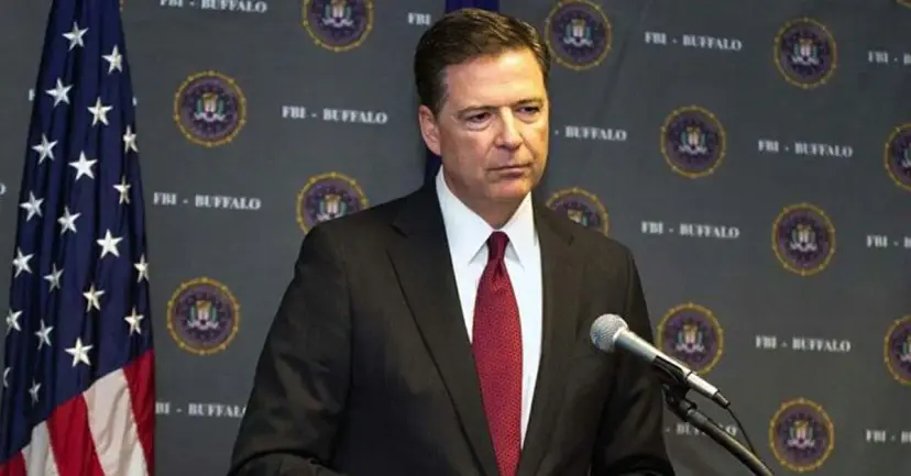 James Comey in a dark suit and red tie speaking at a microphone before an FBI backdrop and American flag.
