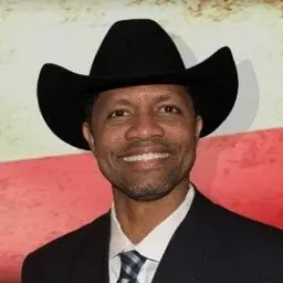 Portrait of a smiling man in a black cowboy hat and dark suit against a red and white background.