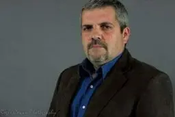 Portrait of a man with graying hair and a goatee wearing a brown jacket over a blue shirt.