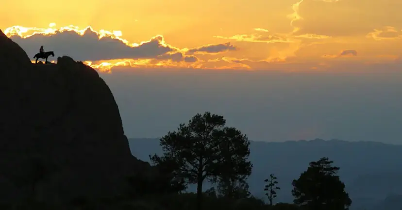 Silhouette of a cowboy on horseback atop a rocky ridge at sunset above a wide valley.