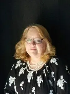 Portrait of a woman with curly blond hair and glasses wearing pearls and a black floral top.