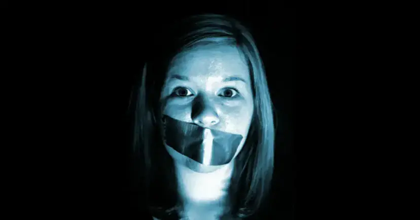 A young woman with tape over her mouth, lit in blue against a black background.