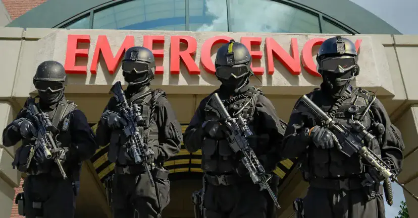 Four armed tactical officers in black gear and helmets standing in front of a hospital emergency entrance.