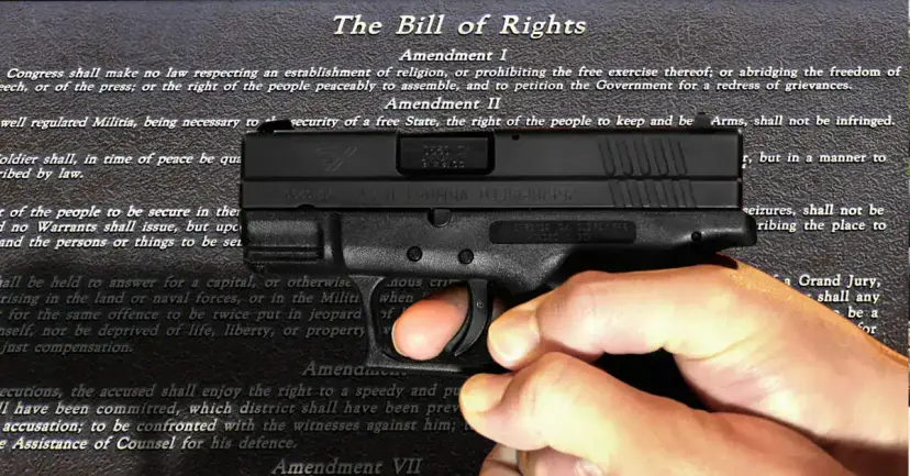 A hand holding a black semiautomatic pistol in front of the printed text of the Bill of Rights.