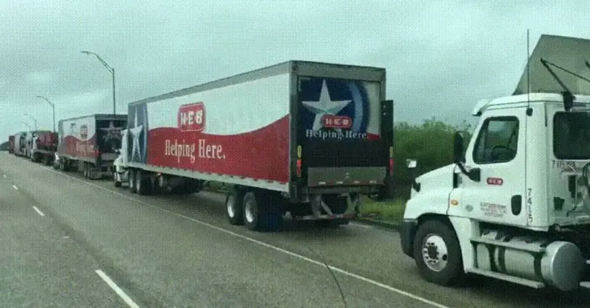 A line of HEB tractor trailers reading 'Helping Here' parked along a highway under gray skies.