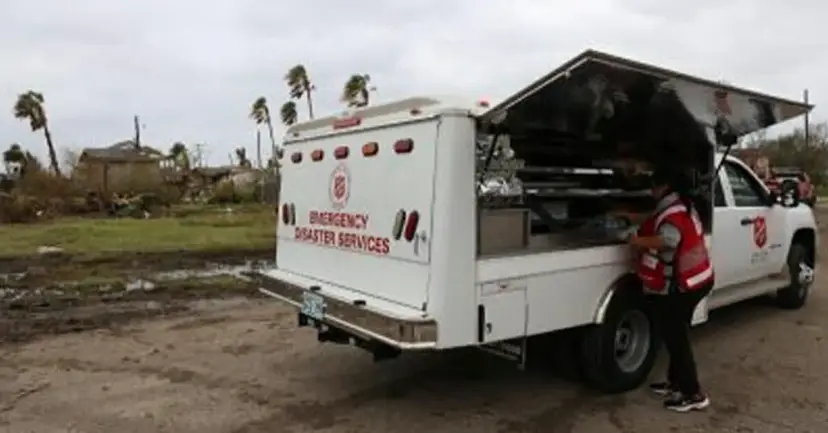 Salvation Army Emergency Disaster Services truck with a volunteer beside it amid storm damage and bent palm trees.