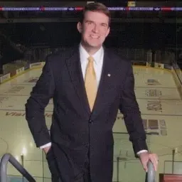 A man in a dark suit and gold tie standing at the rail above an ice hockey rink.
