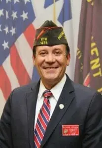 A smiling man in a VFW garrison cap and suit standing in front of flags.