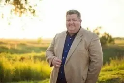 A man in a tan blazer standing in a grassy field at sunset.
