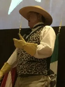 A costumed reenactor in a wide-brimmed hat, leopard-print vest and yellow gloves speaking on stage beside flags.