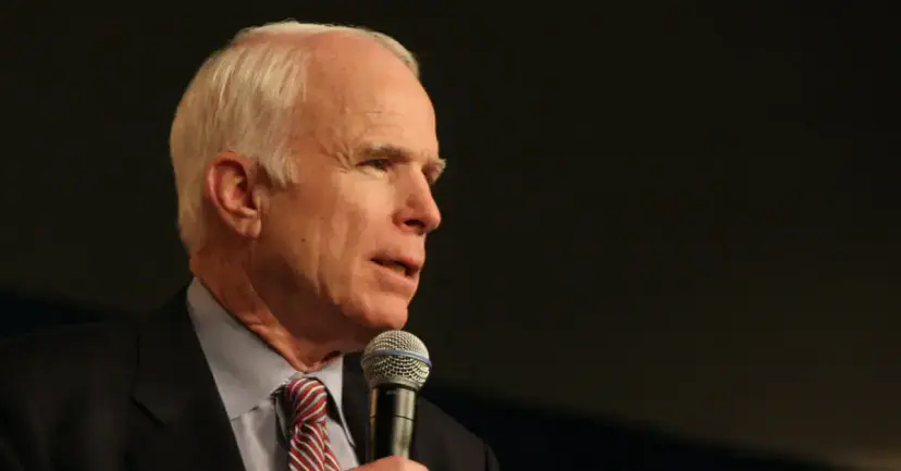 Senator John McCain speaking into a handheld microphone against a dark background.