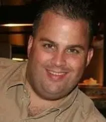 Close photo of a smiling man with short dark hair in a tan shirt.