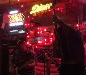 A band playing in a dim bar lit by red lights, with neon Shiner and XX beer signs.
