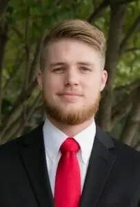 Portrait of a young bearded man in a dark suit and red tie outdoors among trees.