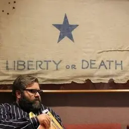 A bearded man in glasses seated beneath a banner with a blue star reading "Liberty or Death."