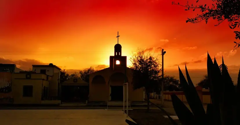 Church bell tower and adobe buildings silhouetted against a fiery orange sunset, with agave plants in front.