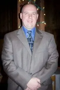 A bald man in a gray suit and blue tie standing with his hands folded indoors.