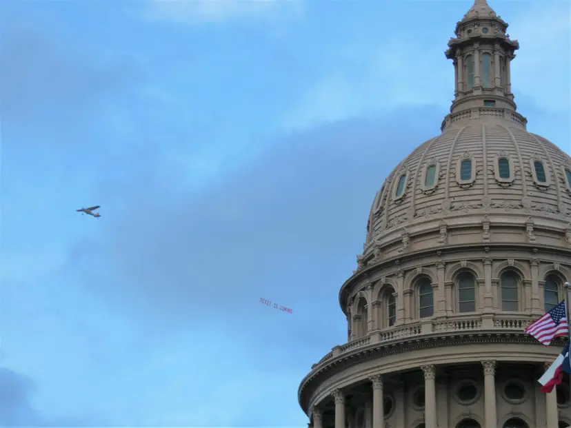 Small plane towing a 'TEXIT is coming' banner past the Texas Capitol dome and its flags.
