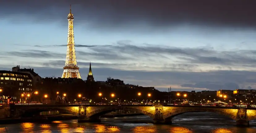 The Eiffel Tower lit up at dusk above the Seine River and a lighted bridge in Paris.