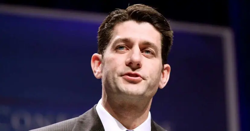 Close portrait of a dark-haired man in a suit speaking against a blue background.